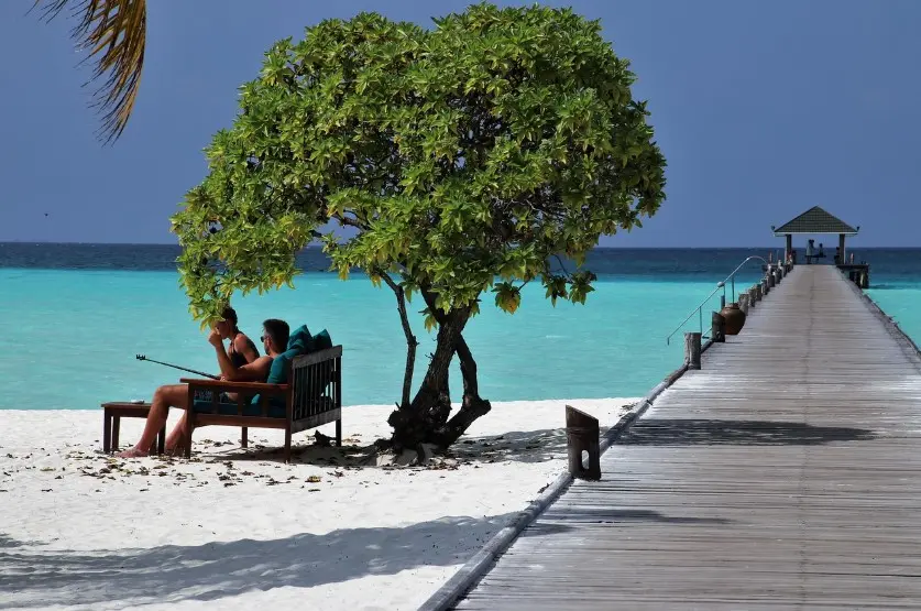 Документы для поездки: что взять с собой в путешествие — пара отдыхает на белом пляже у бирюзового моря, рядом длинный деревянный пирс к беседке.