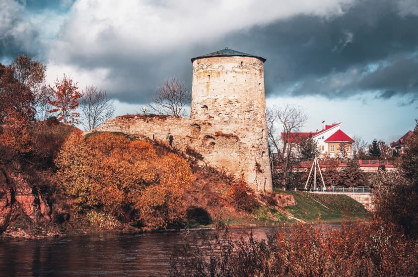 Когда лучше ехать на экскурсию по России: старинная крепостная башня у реки на фоне осенних деревьев