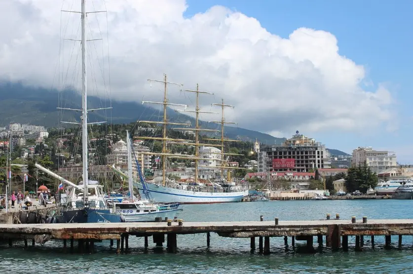 Сколько стоят экскурсии в России: морская набережная с портом, парусником и горами на фоне облачного неба