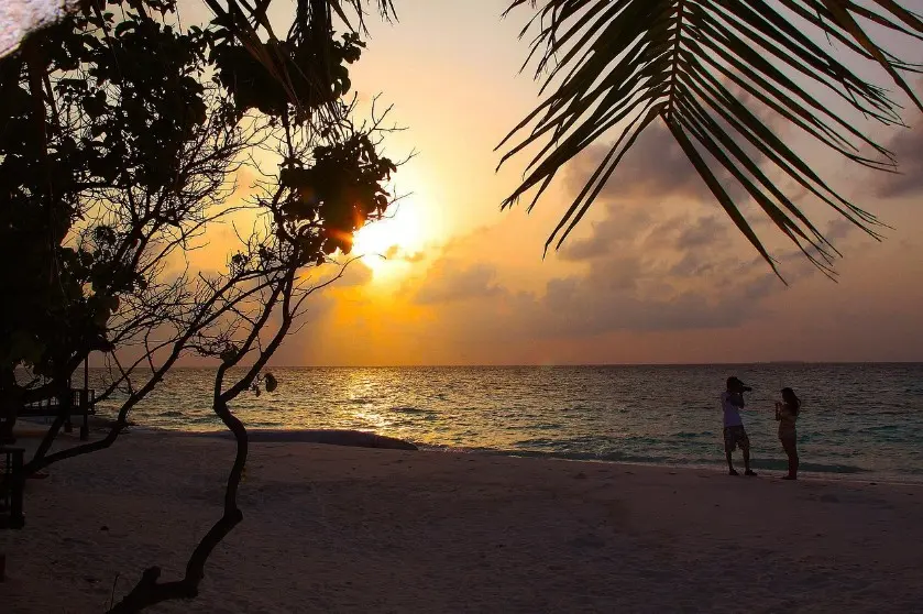 Страховка от невыезда: когда нужна и как работает — закат на пляже у моря, силуэты пары на берегу, пальмы и тёплый свет солнца.